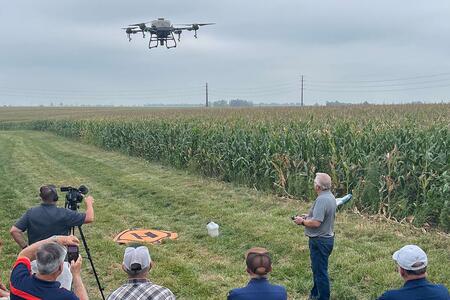A group of people watching an Extension educator demonstrate flying a spray drone near a farm field.