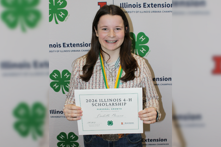 A youth stands in front of a step and repeat banner which shows the 4-H and Illinois Extension logos, holding a certificate
