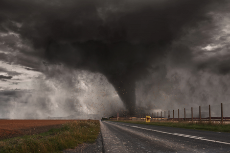 A dark tornado is twisting through rural fields and a two lane road.