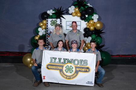 two standing adults and four seated youth holding a banner that says "Illinois Shooting Sports, Learn By Doing"