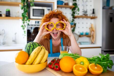 Playful Woman with Vegetables and Fruits, Healthy Diet and Fun Nutrition Concept. Redhead woman in kitchen holding yellow pepper rings over eyes with a variety of fresh produce on the counter.