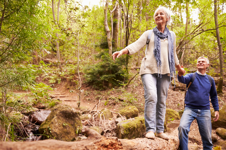 In the woods, a white-haired woman balances on a log while holding a man's hand.