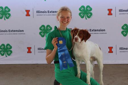 a girl kneels next to a dog and holds a blue ribbon