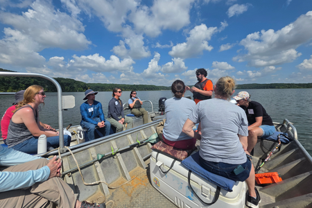 Adults participate in an educational boat tour on a lake under a blue sky.