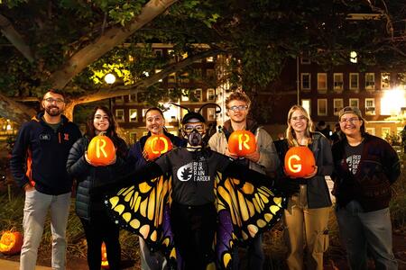 Seven people at night in front of a college residence hall. Four are holding pumpkins. One is dressed like a butterfly.