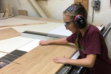 Working under her father Dominic’s watchful eye, Evie Ribbing cross cuts a sheet of veneer core bamboo using a saw.