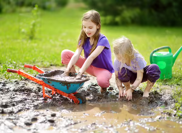 Two funny little girls playing in a large wet mud puddle on sunny summer day. Children getting dirty while digging in muddy soil. Messy games outdoors.