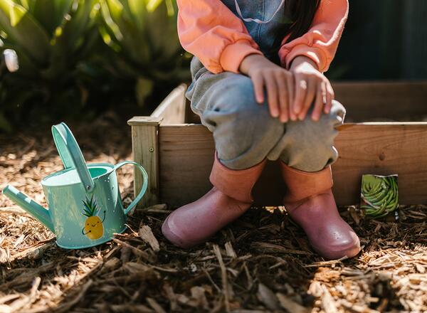 youth in pink boots sitting on raised bed in garden with watering can