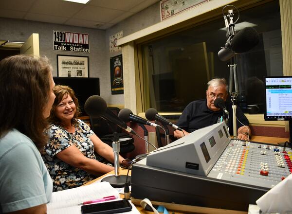 People sitting in a radio studio talking. The on in the middle is smiling.