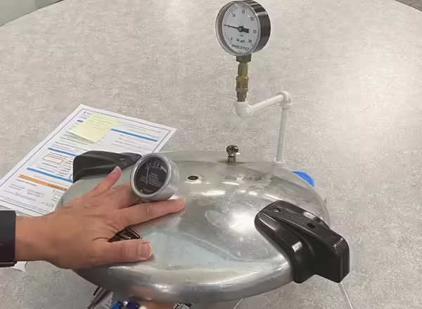 pressure canner lid on table. There's a gauge and it is hooked to some air pipes. The gauge is being tested for accuracy