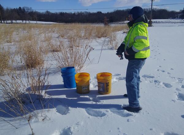 A person standing next to some buckets of seed in a prairie gets ready to sow seed in the snow