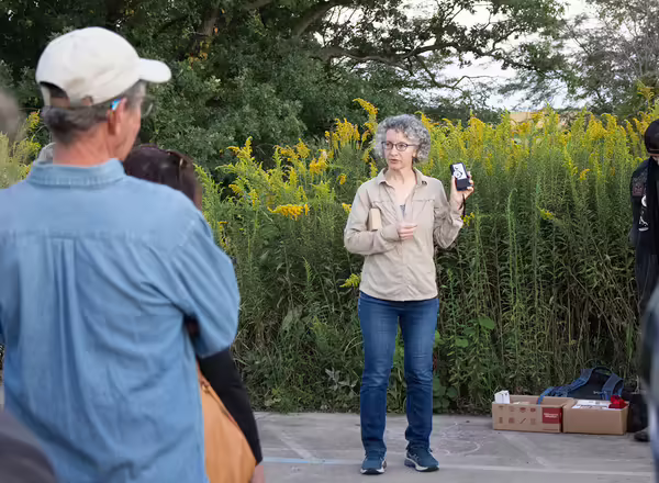 A woman holds a bat sound recorder in front of a crowd at dusk