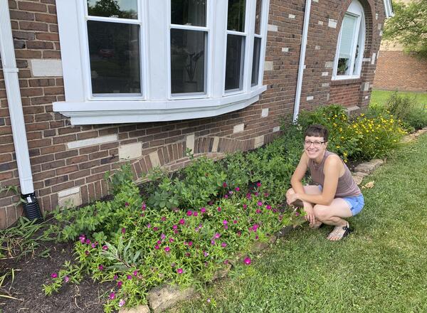 woman with glasses next to flower garden in front of brick building