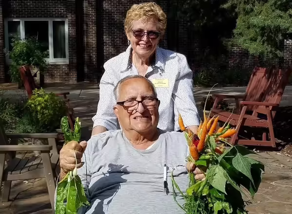 A woman stands behind a man in a wheelchair who holds up produce grown in their garden