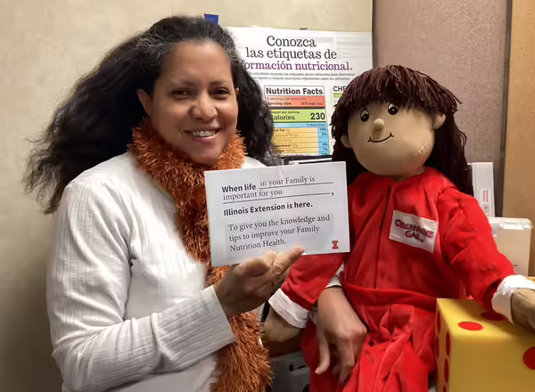 Isabel looks at the camera holding an Eat.Move.Save. doll.  See caption for wording on sign. 