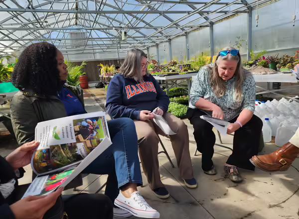 Three people are seated inside a greenhouse reviewing a handout with pictures of horticulture programs. One person has a sweatshirt on that says "Illinois"