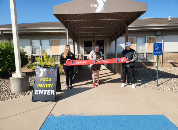 Three people stand in front of a building with a sign that says "YMCA Food Pantry Enter." They are cutting a large, UIUC ceremonial ribbon