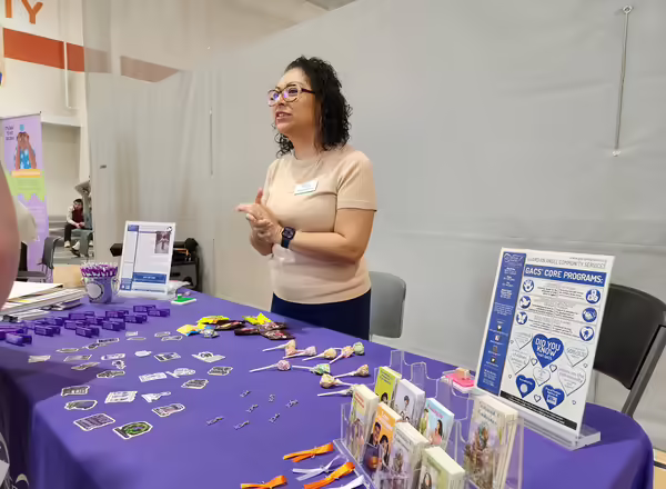 A person stands at a table with a purple tablecloth, speaking to students
