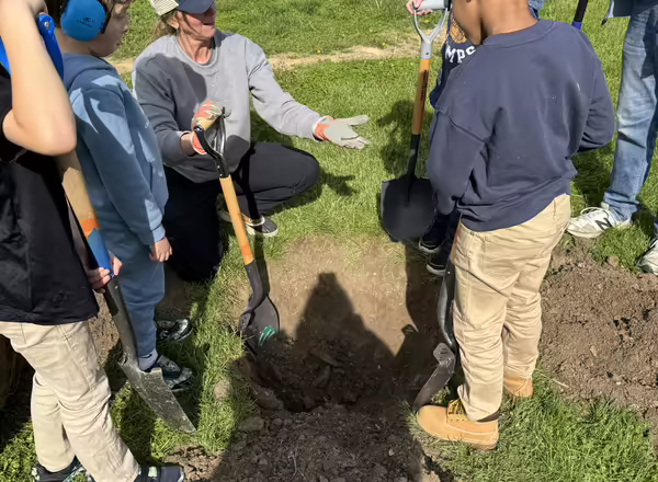 Emily Swihart, horticulture educator kneels with shovel in hand, guiding students in digging a hole to plant thier tree