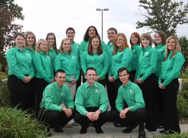 Members of the 4-H youth leadership team pose for a picture. They are all wearing green dress shirts and black pants. Kevin is crouched in the center of the group.
