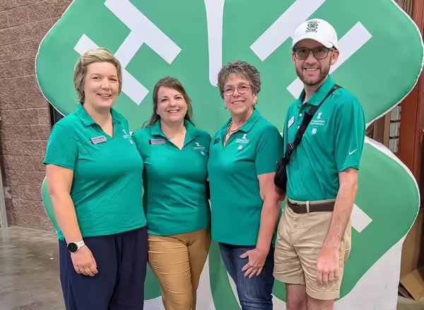 Four 4-H staff members wearing green polo shirts pose in front of a large 4-H Clover. Kevin is on the right side wearing a 4-H baseball cap