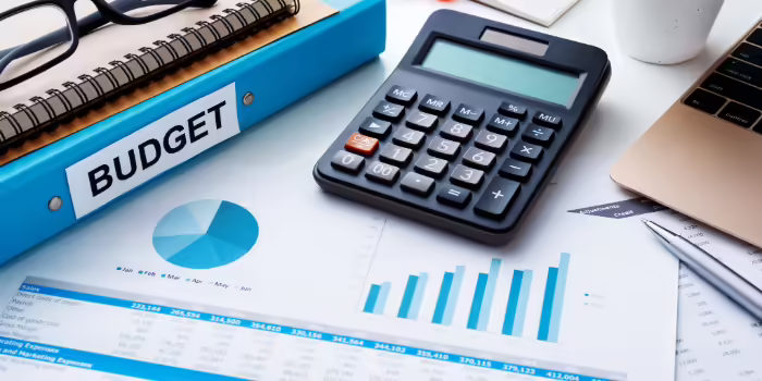 A calculator, budget binder, eyeglasses, and financial charts arranged on a desk beside a laptop and notepad.