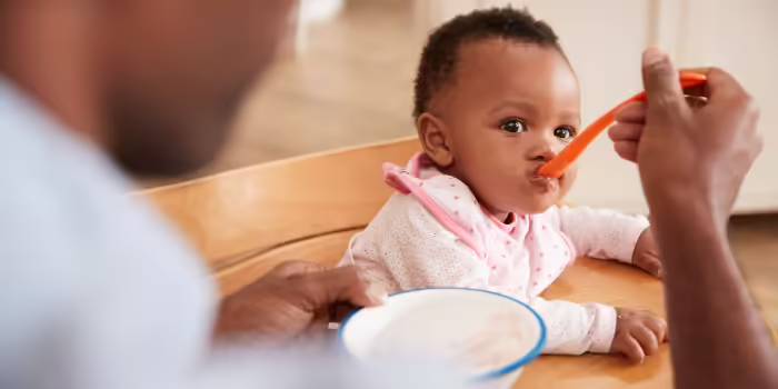 An adult feeding a baby seated in a highchair with a spoon while holding a bowl of food at a dining table.