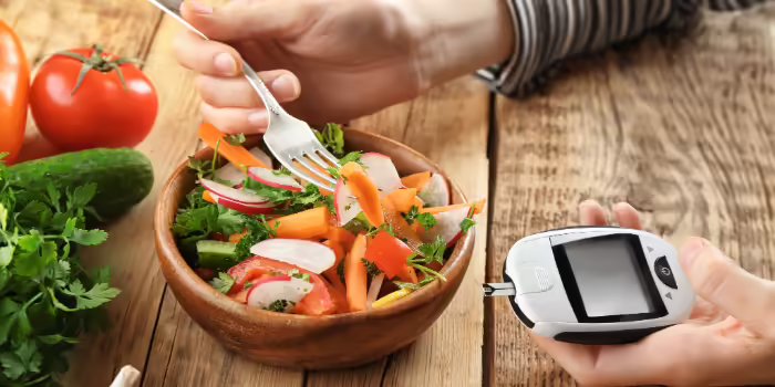 A person eating a colorful vegetable salad while holding a blood glucose meter next to the bowl on a wooden table.
