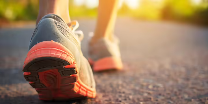 A close-up view of athletic shoes walking on a paved path outdoors, with one shoe in sharp focus and the other blurred in the background. 