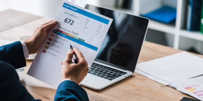 A person reviewing a printed credit report showing a credit score while holding a pen at a desk with an open laptop in the background.