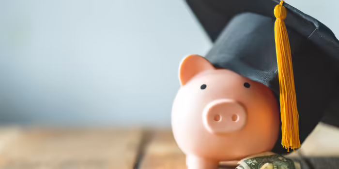 A piggy bank wearing a graduation cap surrounded by dollar bills and coins on a wooden surface.