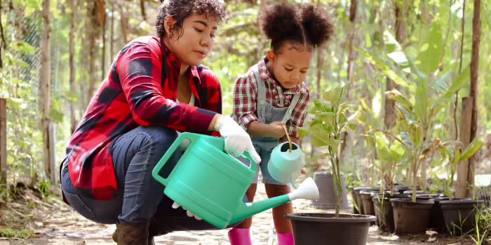 An adult and child watering young potted plants together in an outdoor garden.