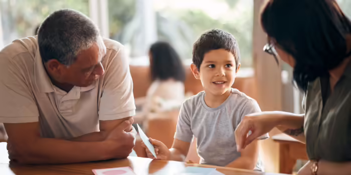 A child holding flashcards while seated at a table with two adults, engaged in a learning activity with educational cards spread across the tabletop.