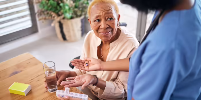An older adult seated at a table with a glass of water while another person offers medication.
