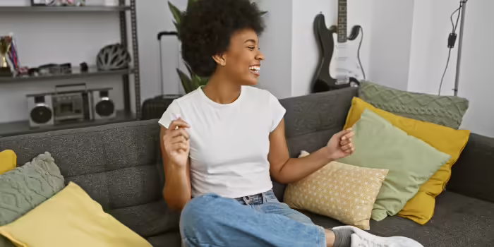 A woman seated on a sofa surrounded by colorful pillows, moving to music in a living room with plants and a guitar in the background.