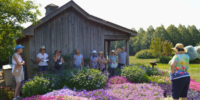 A group standing in front of a shed behind some flowers listening to a speaker