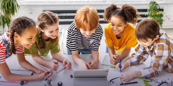five upper elementary age children gathered around a laptop with robotics related items scattered on the table around them