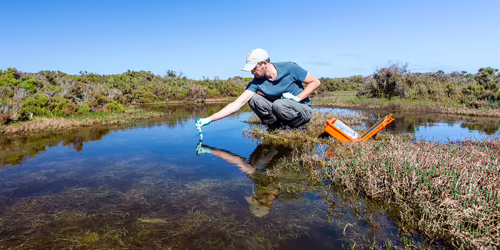 a man crouches next to a pond collecting a water sample 