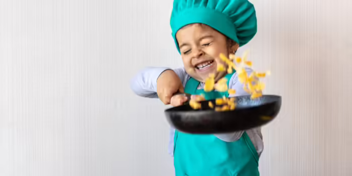 A young child dressed in a teal chef hat and apron smiles joyfully while tossing food in a frying pan, with pieces of pasta flying into the air.