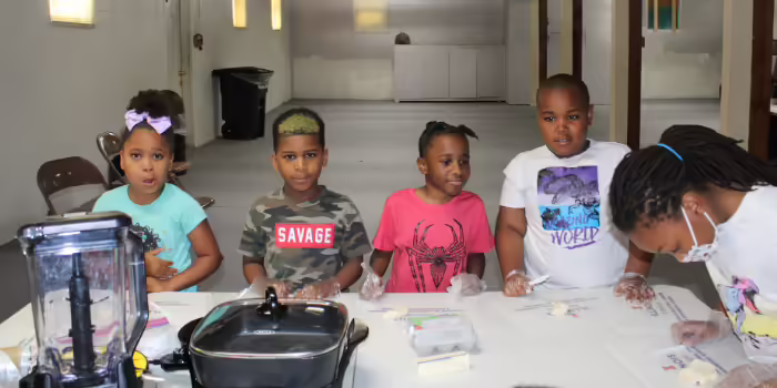 Five children wearing disposable gloves stand and sit around a table during a cooking class.