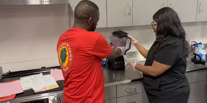 Two participants wearing disposable gloves prepare a fruit smoothie in a teaching kitchen.