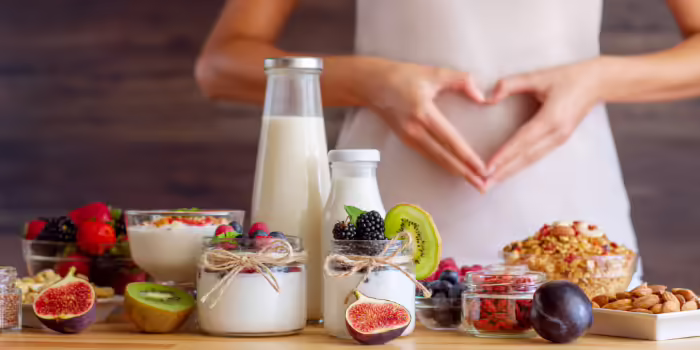 A table with yogurt jars topped with fresh fruits, nuts, and seeds, and there is a person who is forming a heart shape with their hands over their stomach.