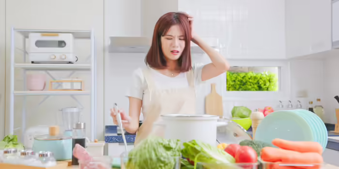 A person standing in a kitchen stirring a pot while looking confused or frustrated, surrounded by fresh vegetables and cooking ingredients on the counter.