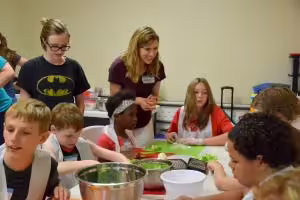 female staff member standing near a group of youth at a table preparing food to make a recipe