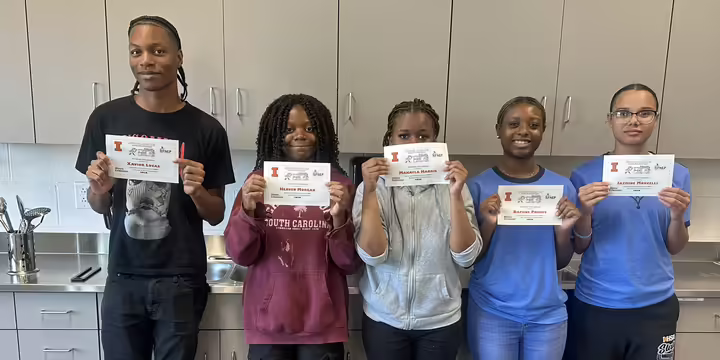 Five youth stand in a classroom kitchen holding certificates from an Illinois Extension cooking program.
