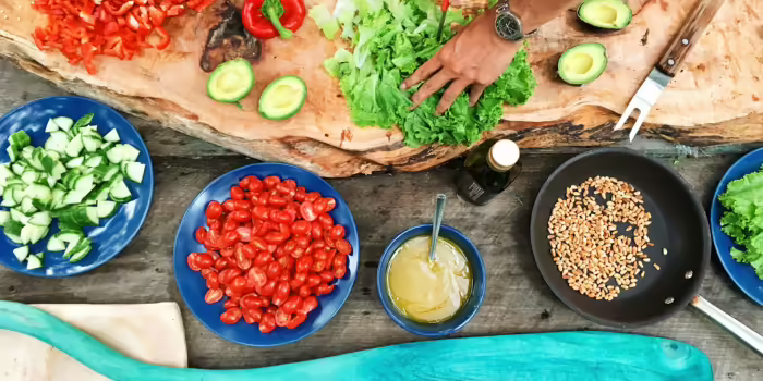 A colorful assortment of food laid out on a table and a wooden board. There is a hand holding down some lettuce to chop it.