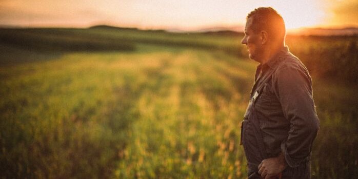 A farmer stands to the right of the image staring out across his fields.