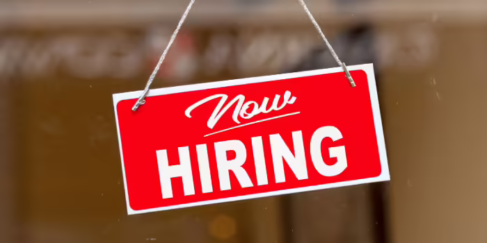 A red and white “Now Hiring” sign hanging by a string in a storefront window, with a blurred business entrance visible in the background.