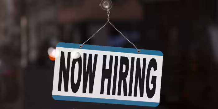A “Now Hiring” sign with bold black lettering on a white background, bordered in blue, hanging by a chain in a storefront window.