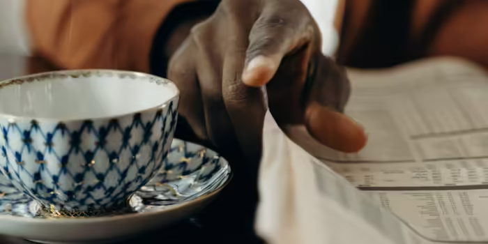 A tea cup next to a hand that is turning the page in a newspaper